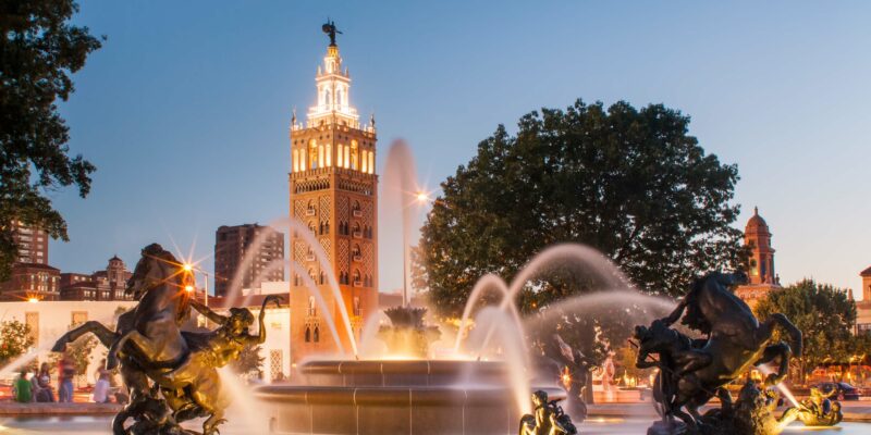 The image shows a fountain with intricate horse sculptures in a lively urban square at dusk. The area features well-lit buildings, including a tall tower with a clock and statues on top, and is surrounded by large trees and city lights, giving a vibrant and picturesque vibe.