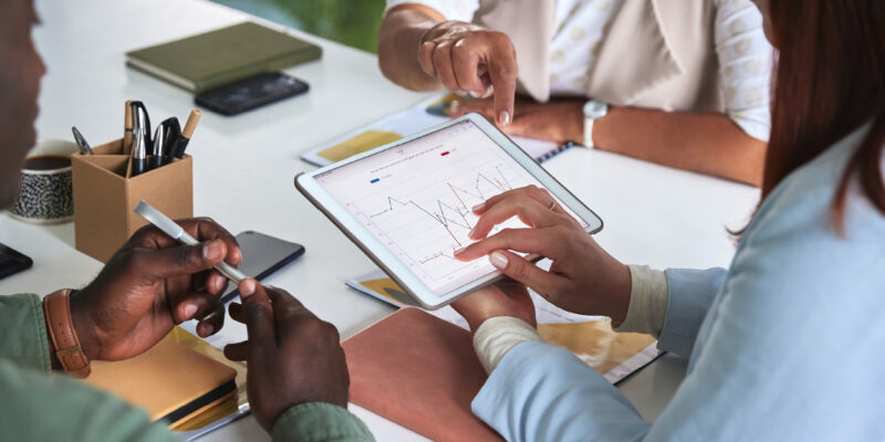 People sitting at a table reviewing a graph on a tablet. One person is pointing at the screen while others hold pens, with notebooks and a coffee cup nearby. The focus is on collaboration and data analysis.