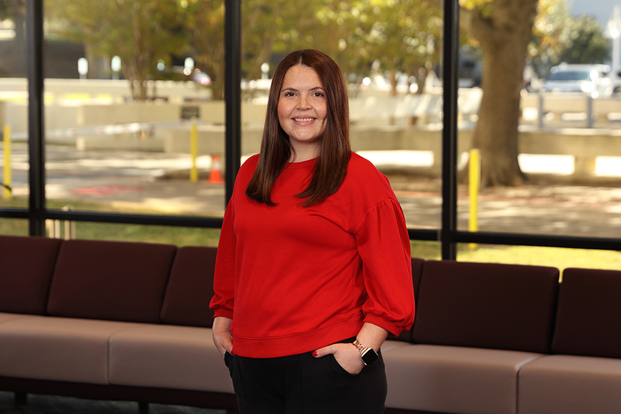A woman with long brown hair wearing a red sweater stands smiling indoors in front of large windows, with a maroon sofa and trees visible outside in the background.