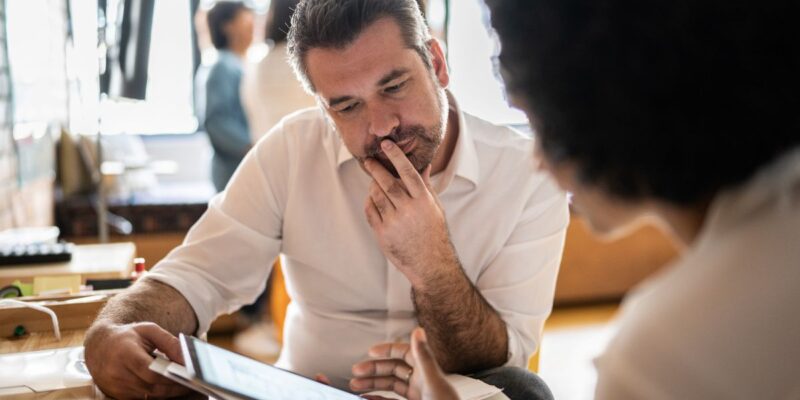 A man in a white shirt looks thoughtfully at a tablet screen held by another person during a discussion in a bright office setting.